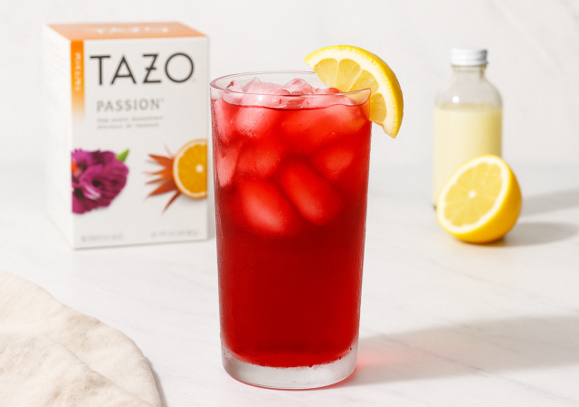 Glass of vibrant red Passion Tea Lemonade with ice and lemon wedge on white marble, with Tazo tea box, halved lemon, and vanilla bottle in soft-focus background.