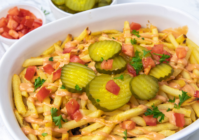 side view of a cheeseburger casserole topped with pickles in a baking dish with condiments around it