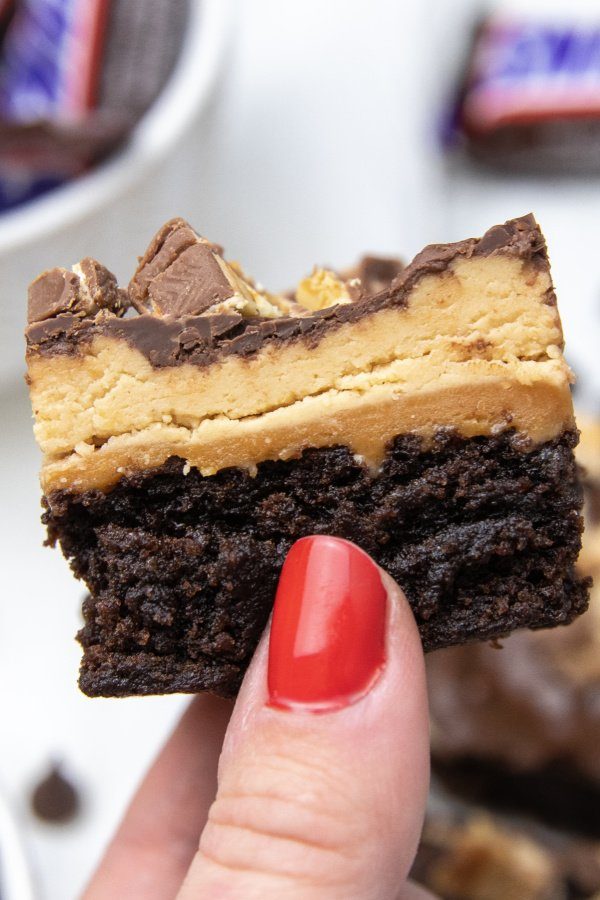 Snicker brownies are a delicious and easy layered brownie. A brownie base, caramel layer, nougat, and chocolate and chopped Snickers! #passion4savings #snickers #brownies #desserttrend #trending #recipe #best #partyfood holding a brownie with snickers layer topping