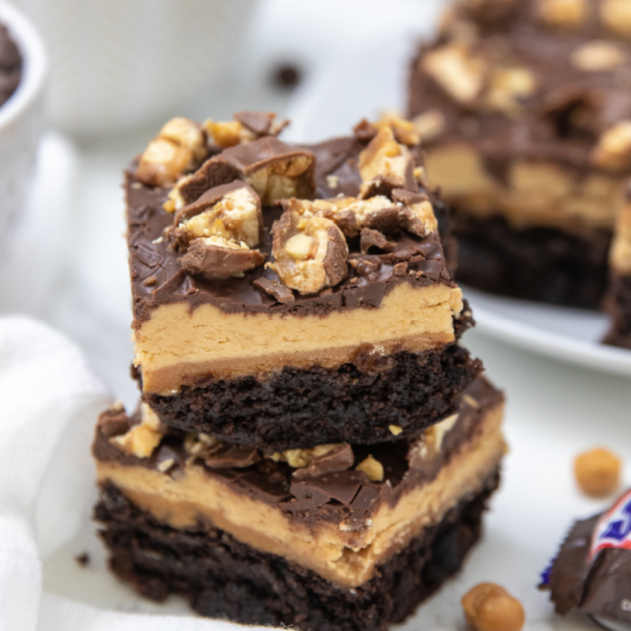 Snicker brownies are a delicious and easy layered brownie. A brownie base, caramel layer, nougat, and chocolate and chopped Snickers! #passion4savings #snickers #brownies #desserttrend #trending #recipe #best #partyfood stacked brownies on white counter