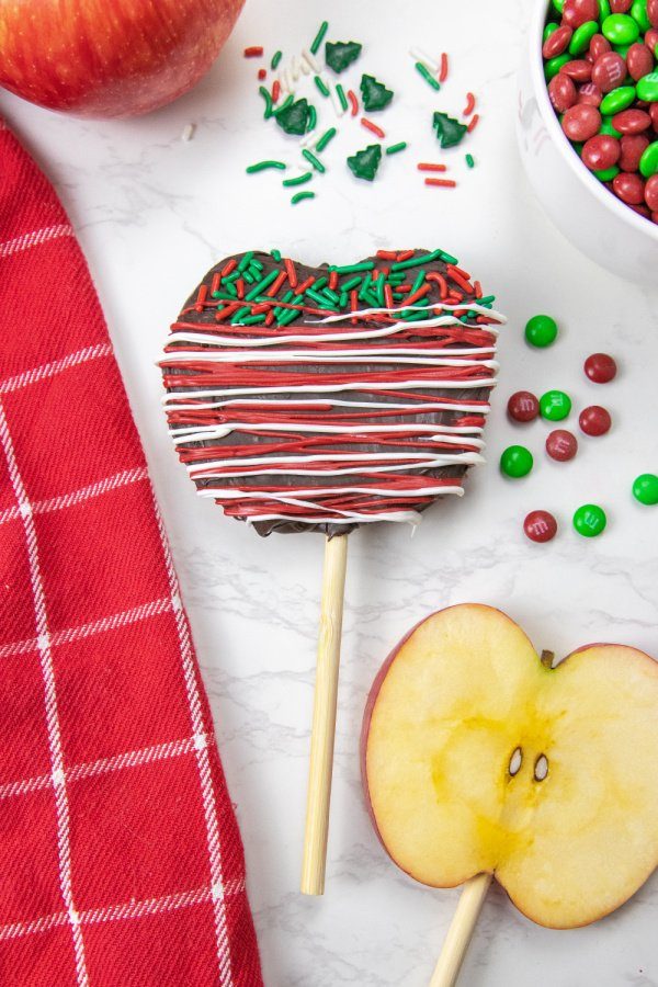 Christmas Chocolate Covered Apple Slices are a simple and easy no bake Christmas treat. Sliced apples decorated and ready to eat in minutes. #passion4savings #apples #dessert #christmas #sliced #dipped #chocolate #chocolatecovered #nobake #kidfriendly apple dipped in chocolate on counter with items around it
