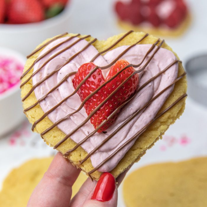 Whip up these quick and easy Mini Valentine Dessert Pizzas. A sugar cookie pizza dessert to serve up this Valentine's Day. #valentinesday #dessert #pizza #holiday #passion4savings #easy #quick #sugarcookie #cookiedough holding heart shaped cookie that is frosted
