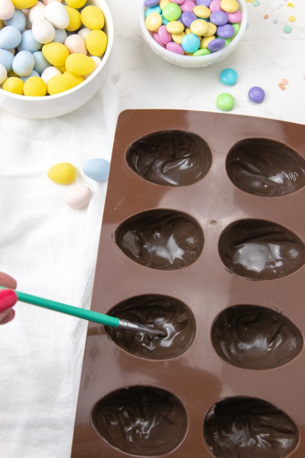 Loaded Cheesecake Easter Eggs are a chocolate candy egg filled with cheesecake and topped with Easter candy. An easy make ahead Easter treat. These fun Easter egg desserts will steal the show. #passion4savings #easter #eggs #dessert #nobake #cheesecake #howto #makeahead painting chocolate into a silicone mold