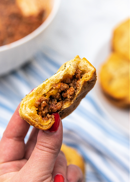 a hand with painted nails is holding a cheesy sloppy joe bomb with a bite taken out of it