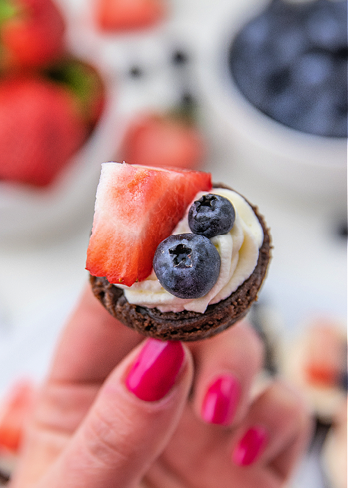 4th of July Brownie Bites are a colorful dessert for all your patriotic parties! Red, white, and blue with strawberries and blueberries! a hand with pink nails holding a patriotic brownie bite