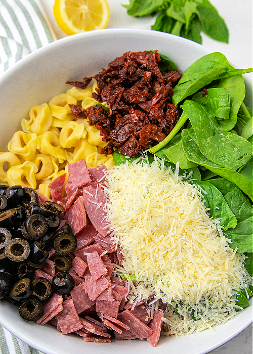 Tuscan Tortellini Salad combines baby spinach, tortellini, salami, black olives, sun-dried tomatoes and more- a light, delicious summer meal! all ingredients unmixed in a white mixing bowl for tuscan tortellini salad