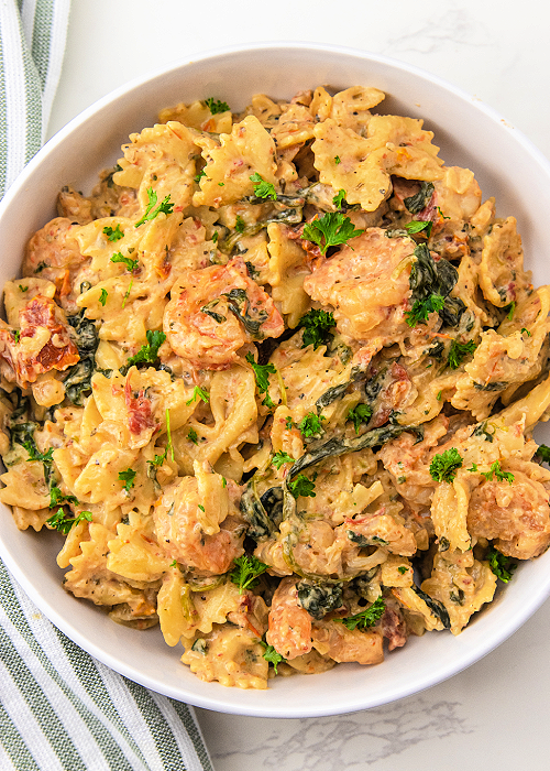 Creamy Garlic Butter Tuscan Shrimp Pasta is a delicious one-pan meal that is healthy, too! Sun-dried tomatoes, parmesan, garlic, and herbs combine for impeccable flavor! garlic shrimp pasta in a white bowl with a green striped napkin